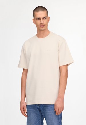 Man with short hair wearing a beige oversized T-shirt and blue jeans, standing against a white background, looking left.