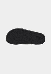 Semelle de chaussure en caoutchouc noir avec motif en vagues texturées, comportant un logo circulaire et des bords renforcés pour une durabilité accrue.