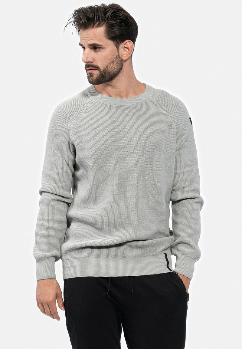 Pull en maille côtelée gris clair avec un col rond et des manches raglan. Présente un accent noir sur la manche gauche et des poignets ajustés.