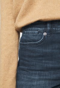 Suéter de punto beige con una textura suave, combinado con jeans de denim azul oscuro con un diseño clásico de cinco bolsillos y detalles de botones de metal.