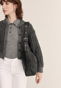 Žena s vlasmi po pleciach, ktorá má na sebe strieborné náušnice, sivý zapínací sveter, biele nohavice a nesie čiernu koženú kabelku cez rameno.