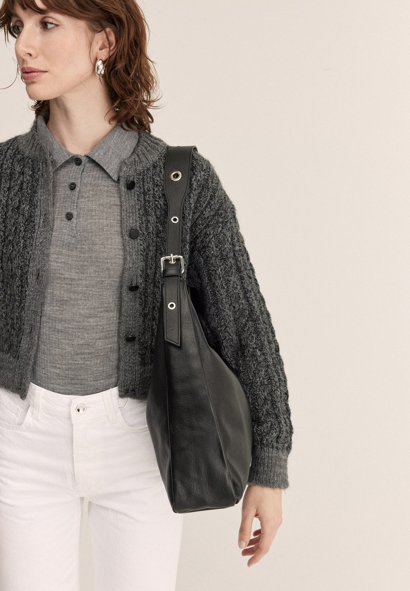 Žena s vlasmi po pleciach, ktorá má na sebe strieborné náušnice, sivý zapínací sveter, biele nohavice a nesie čiernu koženú kabelku cez rameno.