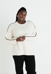 Κρέμα πουλόβερ με πλέξιμο ριμπ και μαύρες ρίγες κατά μήκος των μανικιών, στρογγυλή λαιμόκοψη και χαλαρή εφαρμογή, σε συνδυασμό με μαύρο παντελόνι.