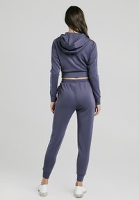 Fialová krátká mikina s kapucí a odpovídající tepláky z měkké látky. Má boční pruhy s barevným přechodem a žebrované manžety.