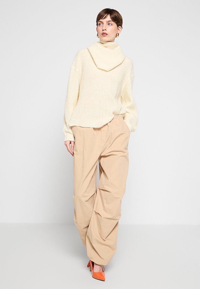 Crème gebreide oversized trui met een col, gecombineerd met beige wijde broek en oranje hoge hakken. Eenvoudige texturen en kleuren.