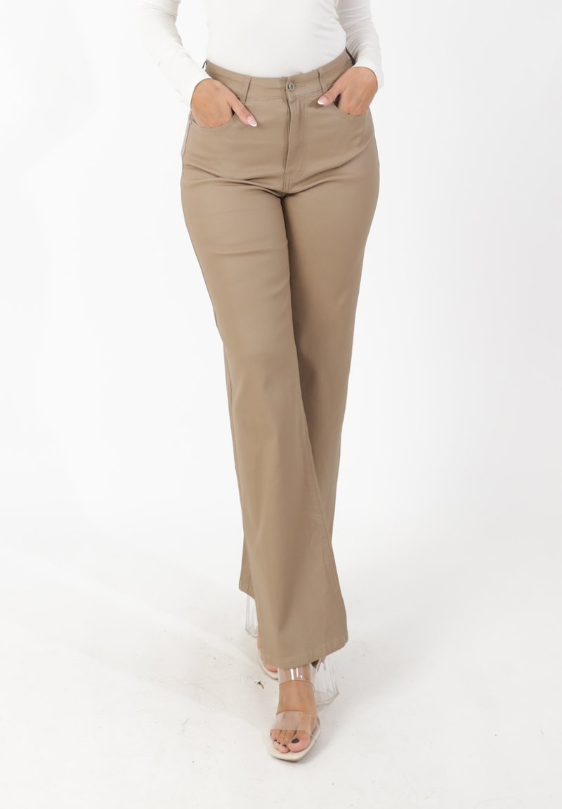 Lichte beige wijde broek met hoge taille, twee voorzakken en een gladde textuur. Draagt bijpassende doorzichtige sandalen met bandjes, gefotografeerd tegen een effen achtergrond.