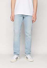 Helle blaue Denim-Jeans haben einen geraden Schnitt mit dezenter Ausbleichung und werden mit weißen Sneakers mit schwarzen Streifen kombiniert. Das Hemd ist schlicht weiß.