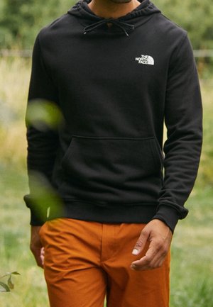 Osoba ubrana w czarną bluzę z kapturem The North Face i pomarańczowe spodnie stojąca na zewnątrz na tle zieleni.