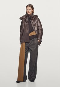 Brązowa kurtka puchowa o błyszczącej powierzchni, noszona na ciemnym swetrze, w połączeniu z szarymi spodniami o szerokich nogawkach z prążkowanym brązowym paskiem.