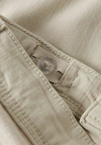 Beige stoff nærbilde som viser en knappestenging og sømdetaljer, med et mykt, strukturert vevmønster.