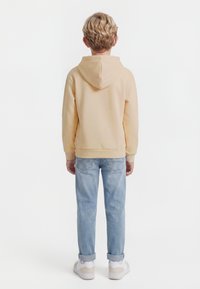 Blonda bērna attēls no muguras, ģērbies bēša krāsas kapučjakā, gaiši zilās sarullētās džinsās un baltos sporta apavos uz vienveidīga fona.