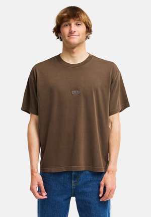 Giovane uomo con capelli castano chiaro che indossa una t-shirt marrone a vestibilità larga e jeans blu, in piedi contro uno sfondo bianco semplice.