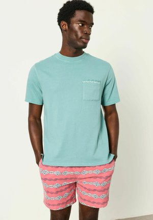 Mann trägt ein hellblaues kurzärmeliges Hemd mit Tasche und pinkfarbene Badeshorts mit horizontalem Fisch- und Wellenmuster.