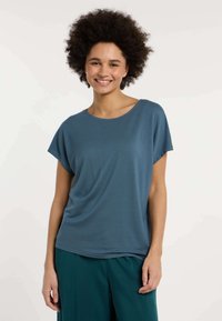 Top blu con maniche corte, dalla vestibilità ampia e rilassata e con una texture morbida; abbinato a pantaloni ampi color turchese, entrambi su uno sfondo chiaro.