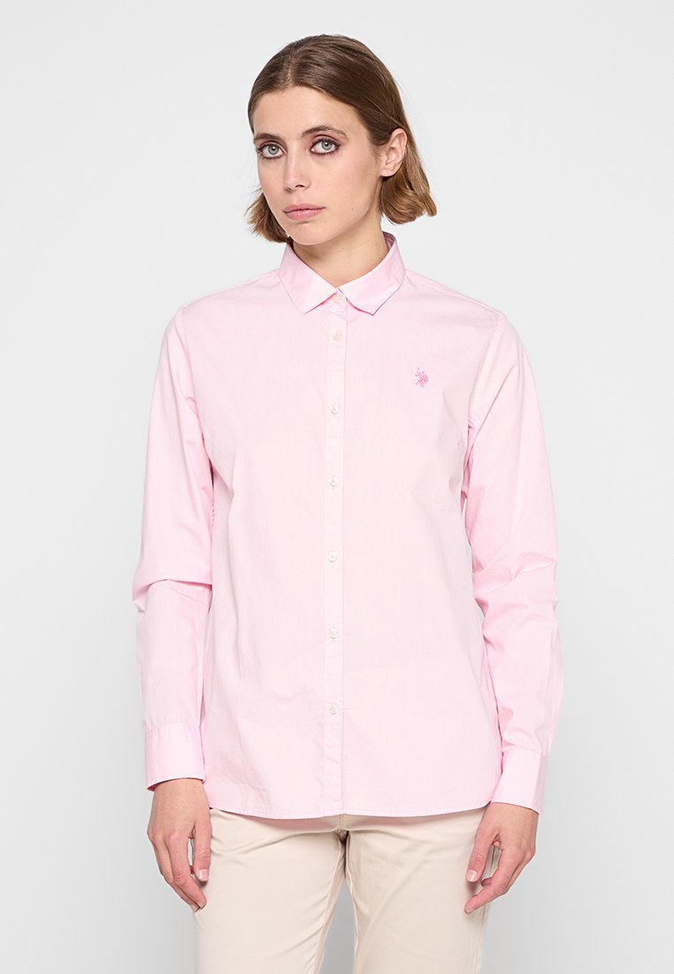 Femme aux cheveux bruns courts portant une chemise à manches longues boutonnière rose clair et un pantalon beige, se tenant devant un fond blanc uni.