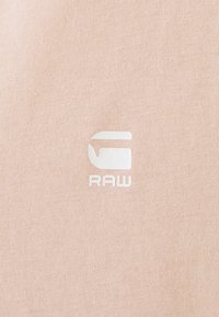Ljusrosa tyg med en slät textur; en vit logotyp präglad i nedre högra hörnet som lyder "RAW" i modern stil.