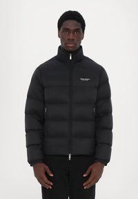 Black puffer jacket with high collar, lightweight, quilted texture, front zip closure, and side zip pockets. Logo on chest.