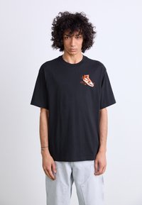 T-shirt en coton noir avec une coupe oversized, présentant un motif de sneaker orange et blanc sur la zone gauche de la poitrine. Manches courtes et col rond.