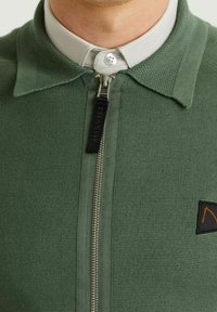 Grön zip-up skjorta i en texturerad stickad kvalitet med vit krage. Har en svart etikett och ett litet orange emblem på bröstet.