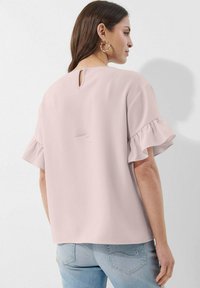 Helles rosa Top mit kurzen Rüschenschultern, Schlüsselaugen-Rückendetail und weichem, glattem Stoff. Getragen mit hellblauen, figurbetonten Jeans.