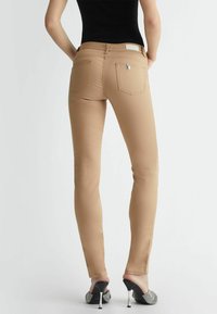 Jeans skinny beige con texture liscia, dotati di tasche posteriori e un dettaglio metallico sulla vita, abbinati a scarpe nere con tacco.