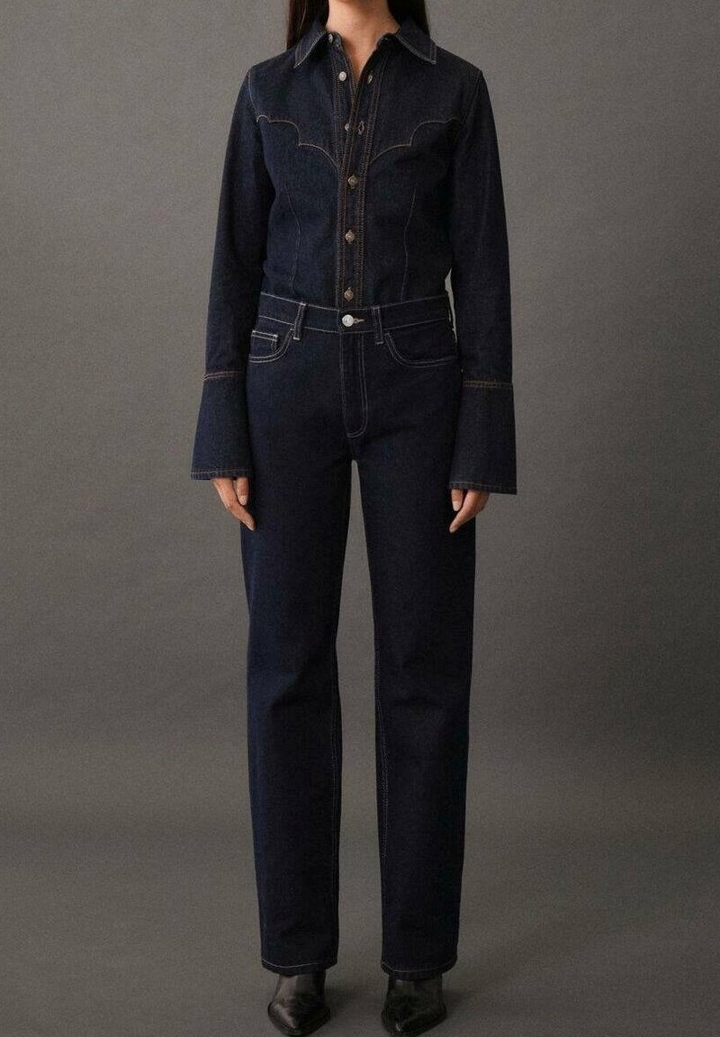 Femme portant une chemise en denim bleu foncé à boutons avec des poignets évasés et un jean droit taille haute assorti sur fond gris uni.