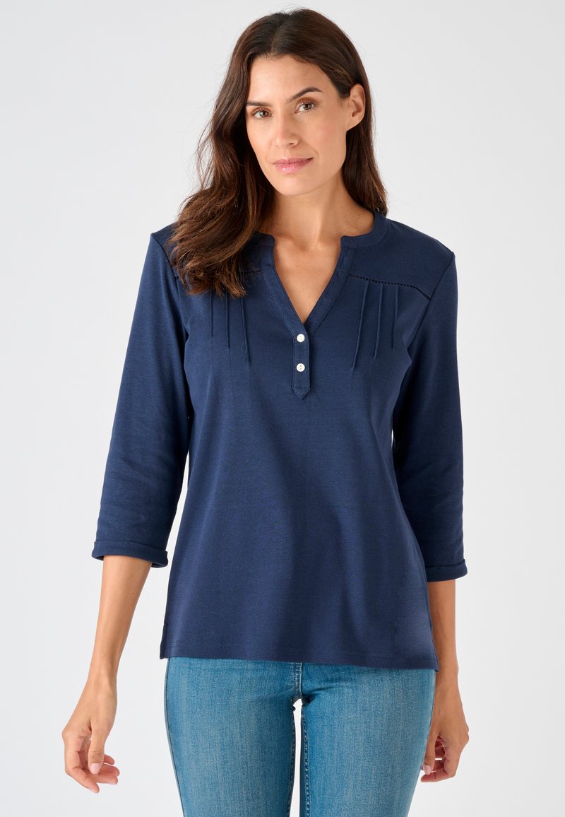 Frau trägt ein marineblaues Shirt mit 3/4-Ärmeln, zwei weißen Knöpfen und dezenten Falten nahe dem Ausschnitt, kombiniert mit hellblauen Jeans.