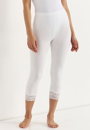 Vrouw draagt witte cropped legging met kanten rand aan de zoom, staat blootsvoets tegen een effen achtergrond.