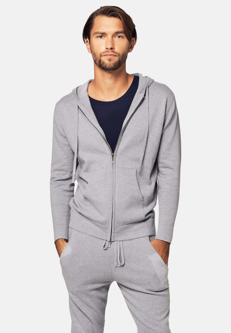 Uomo con barba che indossa una felpa grigia con zip e pantaloni della tuta, in piedi con le mani in tasca su uno sfondo bianco.