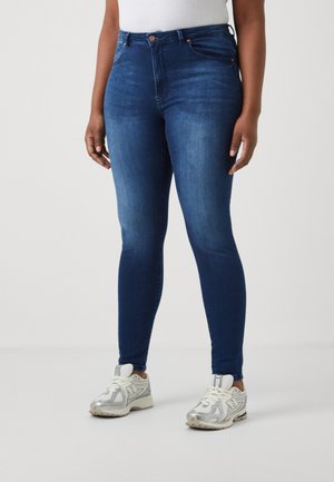 Blugi skinny de culoare albastru închis, cu talie înaltă, având închidere frontală cu button și bucle pentru curea, asortați cu pantofi sport albi și argintii.