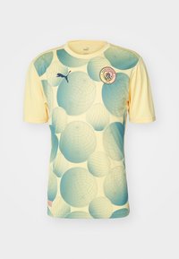 Světle žluté tričko s krátkým rukávem, které má velké tyrkysové a modré kulaté vzory, kulatý límec a vyšitý logo Manchester City.