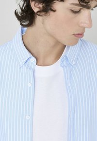 Chemise à rayures bleu clair avec boutons, portée par-dessus un t-shirt blanc à col rond. La chemise possède un col et une texture de tissu douce.