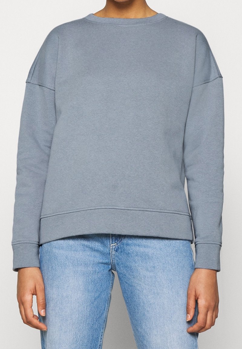 Grå bomullströja med sänkta axlar, ribbade muddar och rundad halsringning. Matchas med ljusblå jeans.