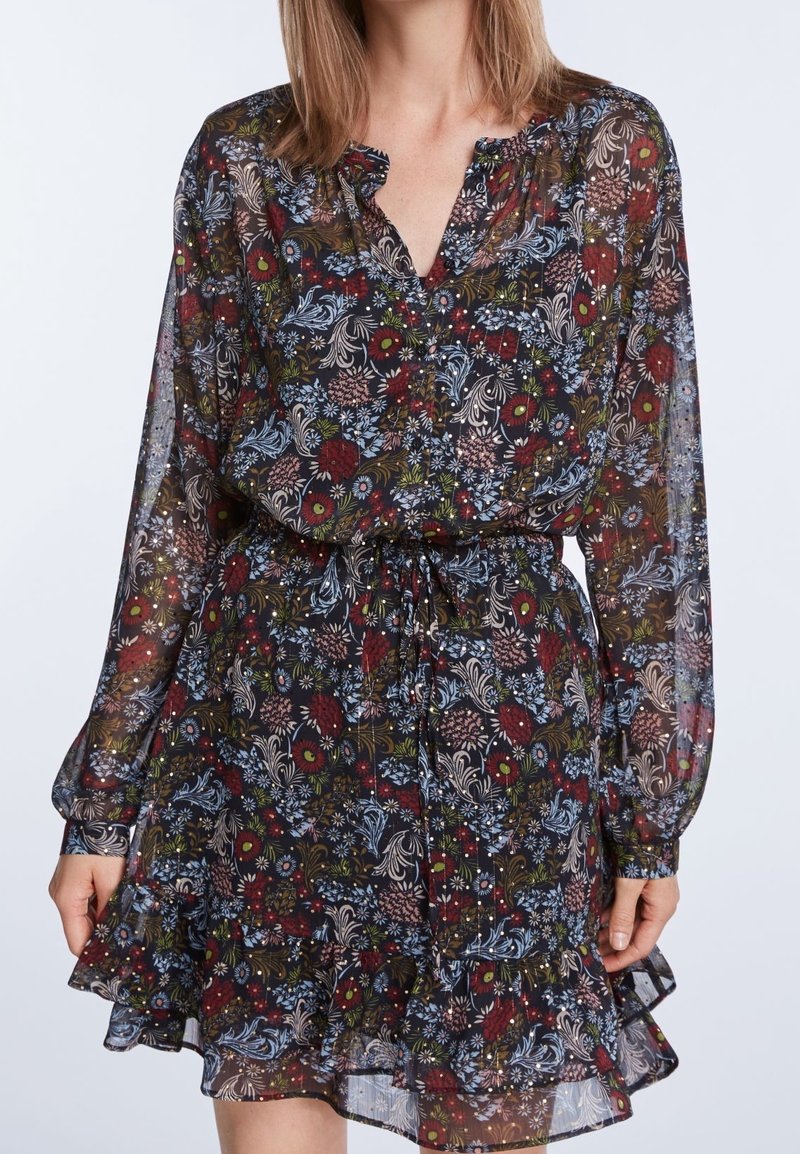 Femme portant une robe fleurie longue transparente à manches longues avec une ceinture à nouer à la taille et un ourlet à volants sur un fond clair uni.