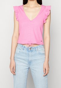 Růžový cropped top s véčkovým výstřihem, volánkovými rukávy a předním zavazováním. Spárováno se světle modrými džíny s vysokým pasem.