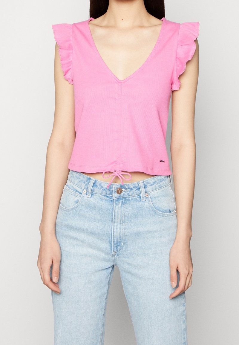 Růžový cropped top s véčkovým výstřihem, volánkovými rukávy a předním zavazováním. Spárováno se světle modrými džíny s vysokým pasem.