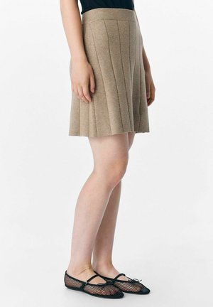 Jupe plissée beige en tissu texturé, dotée d'une taille ajustée et d'une silhouette évasée, associée à des ballerines en mesh noires.