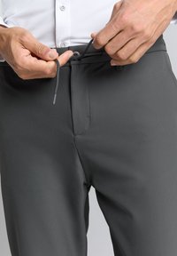 Grijze, lichtgewicht broeken met een gladde textuur, trekkoord en knoopsluiting. Close-up van handen die de tailleband aanpassen.