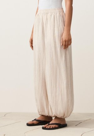 Femeie purtând pantaloni largi bej deschis cu dungi subțiri și manșete elastice, asortați cu șlapi negri pe un podea placată cu gresie.