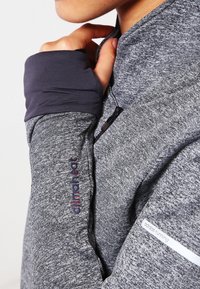 Osoba nosząca szaro-srebrną kurtkę biegową Adidas z technologią Climawarm, trzymająca kołnierz przy brodzie, pokazująca otwór na kciuk w mankiecie rękawa.