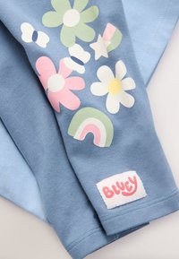 Camiseta de algodón azul con estampados florales coloridos y arcoíris. Incluye una etiqueta blanca con el texto "Bluey." Textura suave, ajuste relajado.