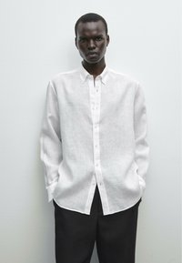 White linen button-up shirt with a relaxed fit, long sleeves, and a classic collar. Paired with dark trousers, set against a minimal background.