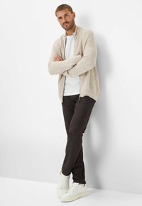 Beige gebreide cardigan met een hoge kraag, gedragen over een wit T-shirt, gecombineerd met donkerbruine broek en witte sneakers. Staande pose, met gekruiste armen.