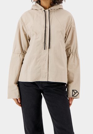 Kobieta nosząca beżową kurtkę z kapturem, czarnymi sznurkami i logo na mankiecie lewego rękawa, w zestawieniu z czarnymi spodniami na jednolitym tle.