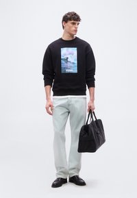 Νεαρός άνδρας που φοράει μαύρο φούτερ με εκτύπωση κυμάτων ωκεανού και κείμενο "HUGO", ανοιχτόχρωμο τζιν, μαύρα παπούτσια, κρατώντας μαύρη τσάντα duffle.