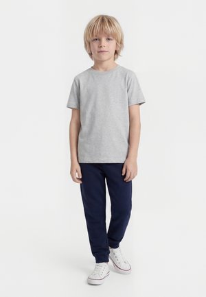 Niño rubio de pie, vestido con una camiseta de manga corta gris claro, pantalones azul oscuro y zapatillas blancas, sobre un fondo blanco liso.