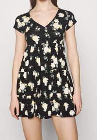 Svart blommig klänning med korta ärmar och V-ringning, prydd med gula rosor och vita accenter, volangkjol, tillverkad av mjukt material.