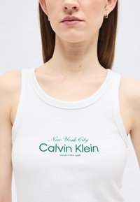 Γυναίκα που φοράει λευκό αμάνικο τοπ με πράσινο κείμενο "New York City Calvin Klein Established 1968" μπροστά, με απλό ανοιχτόφωτο φόντο.