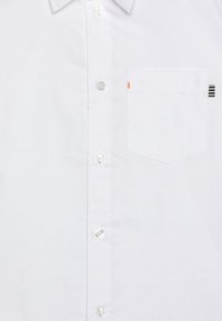 Camisa blanca de botones con un bolsillo en el pecho que tiene un acento naranja y rayas negras, confeccionada en una tela texturizada y con un cuello estructurado.