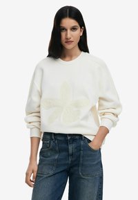 Cream sweatshirt with a textured floral design, featuring ribbed cuffs and a round neckline. Paired with high-waisted, dark blue jeans.